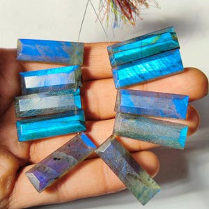 May include: A selection of rectangular labradorite gemstones, exhibiting iridescent blue, teal, and purple tones. The stones are faceted, reflecting light and revealing their internal colours. The gemstones are held in a hand, demonstrating their size and shape.