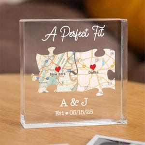 Puede incluir: Bloque acrílico transparente con un diseño de mapa que presenta dos piezas de rompecabezas, una para Nueva York y otra para Dallas, conectadas por una línea. El texto "A Perfect Fit" está en la parte superior, con "A & J" y "Est 05/15/25" en la parte inferior.