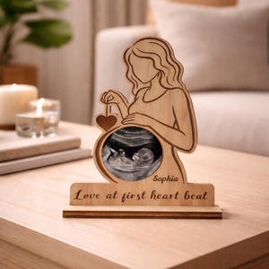 May include: Wooden pregnancy announcement decor. The design features a pregnant person silhouette holding a heart, an ultrasound image, the text "Love at first heart beat", and the name "Sophia".