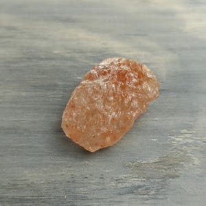 May include: A raw, unpolished sunstone crystal with a translucent, peach-orange hue. The mineral features a rough, uneven surface texture with natural crystalline facets that catch the light, displaying internal shimmer and varied warm color tones.