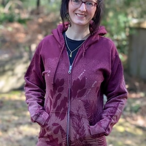 May include: A burgundy zip-up hoodie with a botanical leaf pattern. The hoodie has a faded design and a full-length zipper. The pockets are visible, and the hood is up. The person is wearing a black shirt underneath.