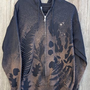 May include: Black zip-up hoodie with a nature-inspired design. The hoodie features a brown and black leaf and fern pattern, with a distressed, speckled effect. The hoodie has a front pocket and a full zipper.