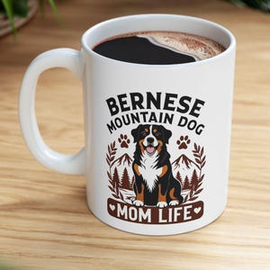 Puede incluir: Una taza de cerámica blanca con un gráfico de un perro de montaña de Berna sentado frente a un paisaje montañoso. El texto dice "BERNESE MOUNTAIN DOG" sobre el perro y "MOM LIFE" debajo, junto a huellas de patas y corazones.