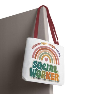 Puede incluir: Un bolso tote blanco con asas de color rojo oscuro que presenta un gráfico de arcoíris colorido sobre un pequeño corazón y el texto SOCIAL WORKER en letras negritas y multicolores. Sobre el arcoíris aparecen las palabras compassion, support, strength, empathy.