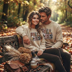 Puede incluir: Dos sudaderas beige de cuello redondo con el texto FALL VIBES en un patrón de estampado de leopardo. Las sudaderas tienen mangas largas y un ajuste relajado. El texto incluye un gráfico de rayo entre las palabras en el frente de cada prenda.