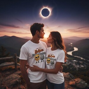 Puede incluir: Dos camisetas blancas con un estampado gráfico de un sol sonriente con gafas de sol y un personaje lunar de pie uno al lado del otro. Sobre los personajes, el texto Total Solar Eclipse aparece en una fuente retro sobre un patrón de cuadros.