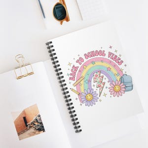 Puede incluir: Un cuaderno de espiral con un gráfico colorido que incluye el texto "BACK TO SCHOOL VIBES" arqueado sobre un arcoíris, un rayo floral, dos flores sonrientes, una regla, un lápiz y una mochila azul sobre una portada blanca.