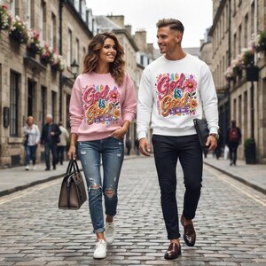 Puede incluir: Una sudadera rosa claro y una sudadera blanca, ambas con el texto gráfico colorido God is Good All The Time con detalles florales. La sudadera rosa se combina con vaqueros azules desgastados y zapatillas blancas. La sudadera blanca se combina con pantalones negros y zapatos de cuero marrón.