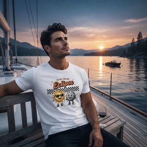 Puede incluir: Una camiseta blanca de manga corta con un gráfico de un sol sonriente con gafas de sol y una luna sonriente, ambos con brazos y piernas. Sobre ellos aparece el texto Total Solar Eclipse sobre un fondo de cuadros blancos y negros.