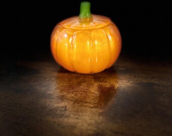May include: A small, round, orange pumpkin-shaped container with a smooth, glossy finish and a distinct ribbed texture. The lid features a short, cylindrical green stem at the top, providing a contrasting color to the vibrant orange body.