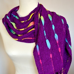 May include: A vibrant purple scarf with colorful ribbon accents and fringe. The scarf is displayed on a mannequin, showcasing its woven texture and the bright hues of the ribbons, including yellow, pink, and blue. The fringe at the bottom adds a decorative touch.