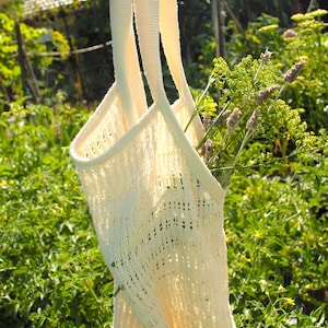 May include: Cream-colored knitted tote bag with long straps, filled with green and purple flowers. The bag has a delicate, open weave design. The background is a lush green garden, with sunlight filtering through.