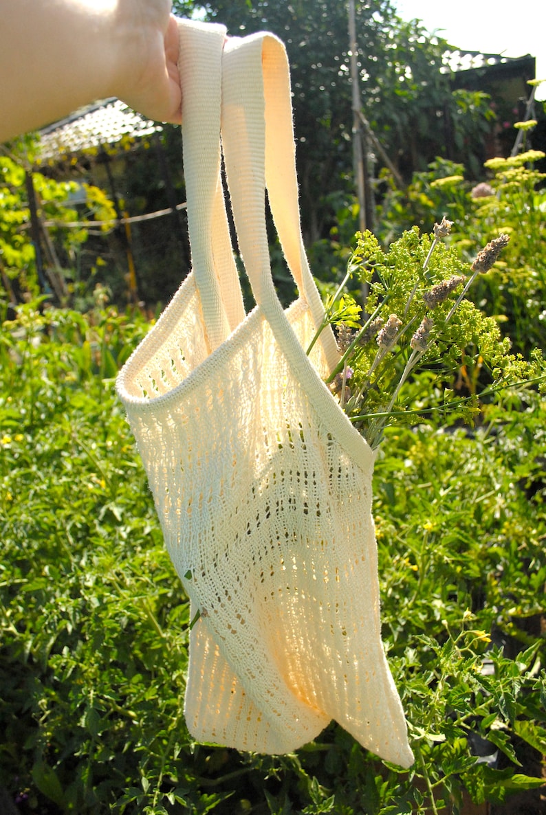May include: Cream-colored knitted tote bag with long straps, filled with green and purple flowers. The bag has a delicate, open weave design. The background is a lush green garden, with sunlight filtering through.