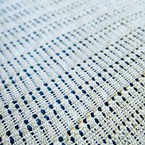 May include: Close-up of a cream-colored, woven textile with a repeating pattern of openwork squares and lines. The fabric has a textured appearance, with a blue background visible through the open weave. The overall effect is delicate and intricate.