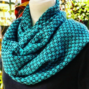 May include: A turquoise blue infinity scarf with a black geometric pattern. The scarf is made of a soft, woven fabric.