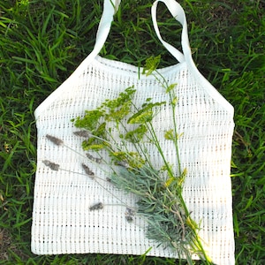 May include: A white, open-weave tote bag with two handles, lying on green grass. The bag is filled with a bundle of lavender and yellow-green wildflowers. The bag's texture is visible, and the overall aesthetic is natural and rustic.
