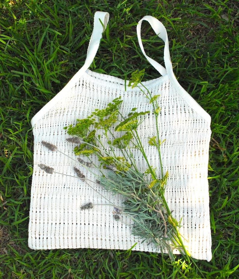 May include: A white, open-weave tote bag with two handles, lying on green grass. The bag is filled with a bundle of lavender and yellow-green wildflowers. The bag's texture is visible, and the overall aesthetic is natural and rustic.