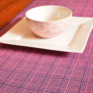 May include: A pink and blue woven table runner with a square pattern. A white square plate and a pink bowl with a white design are on top of the runner.
