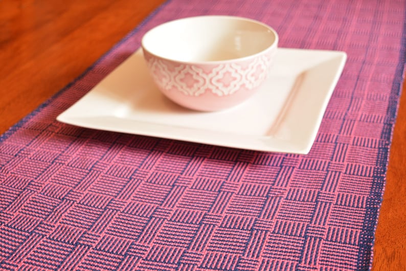 May include: A pink and blue woven table runner with a square pattern. A white square plate and a pink bowl with a white design are on top of the runner.