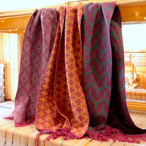 May include: Four woven scarves in different colors and patterns. The scarves are hanging on a wooden rack. The scarves are made of a soft, textured fabric. The scarves are all different lengths.