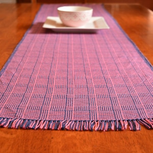 May include: A pink and blue woven table runner with fringe on the ends. The runner is on a wooden table.