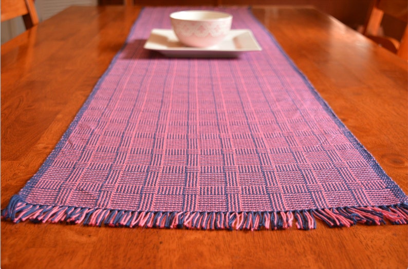May include: A pink and blue woven table runner with fringe on the ends. The runner is on a wooden table.