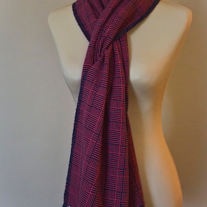 May include: A pink and navy blue patterned scarf with fringe on the ends. The scarf is tied around a mannequin's neck.