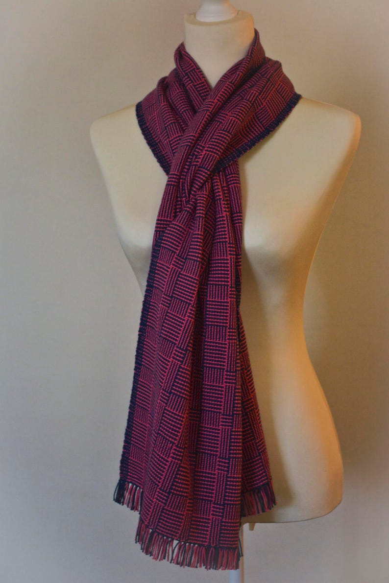 May include: A pink and navy blue patterned scarf with fringe on the ends. The scarf is tied around a mannequin's neck.
