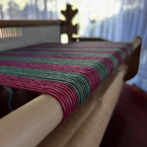 May include: Close-up of a wooden weaving loom with a partially woven textile. The textile features alternating stripes of pink and green yarn. The loom is made of light-colored wood, and the background is blurred.