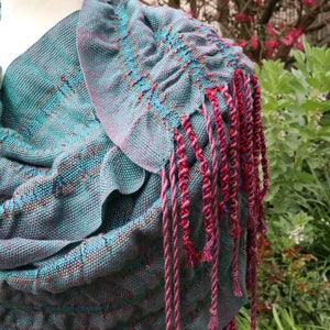 May include: A teal blue woven shawl with a pink and red fringe. The shawl is draped over a mannequin and has a textured woven pattern.