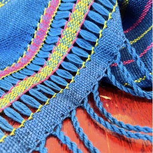 May include: A blue woven fabric with pink and yellow stripes and fringe. The fabric is folded and the fringe is hanging down.