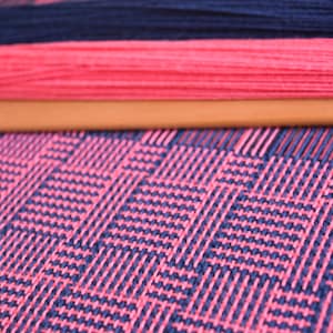 May include: Close-up of a loom with pink and dark blue yarn woven into a checkered pattern.