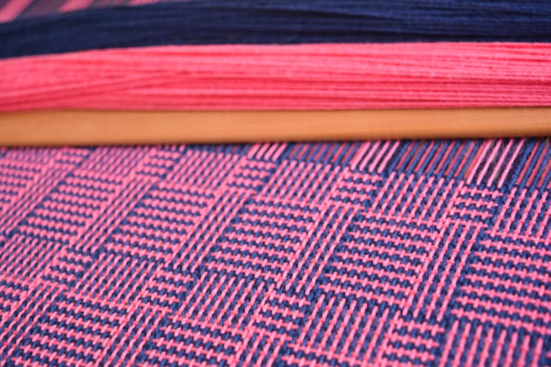 May include: Close-up of a loom with pink and dark blue yarn woven into a checkered pattern.