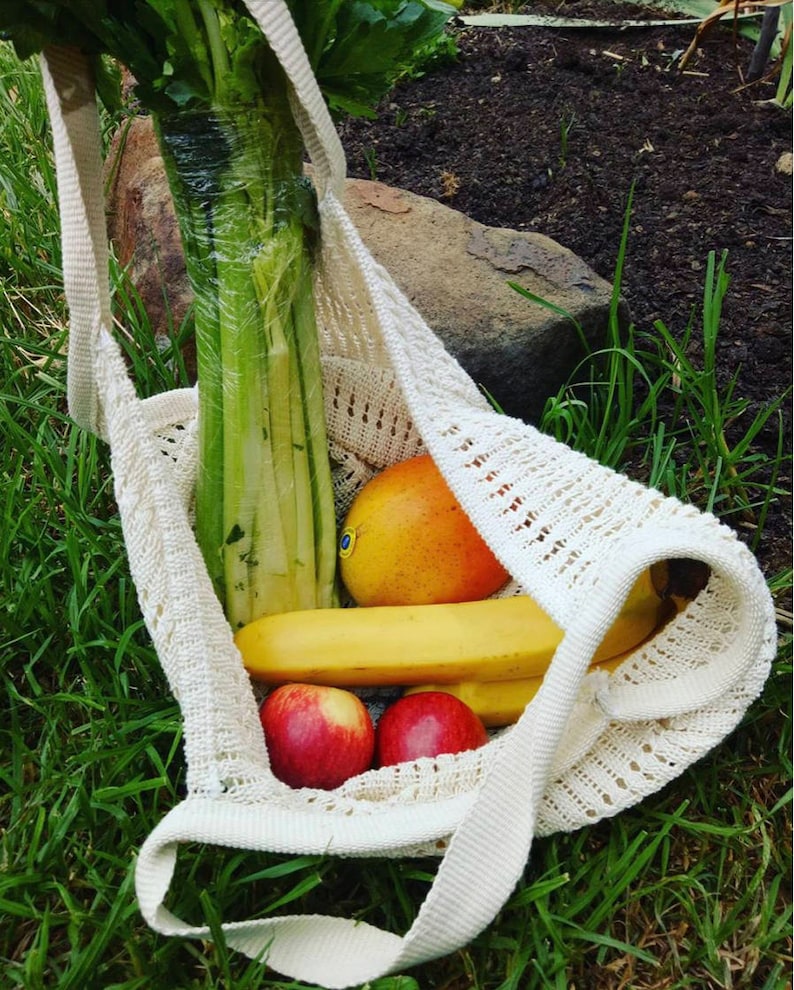May include: A cream-colored, open-weave tote bag filled with fresh produce. The bag contains a bunch of celery, a mango, bananas, and red apples. The bag is sitting on green grass.