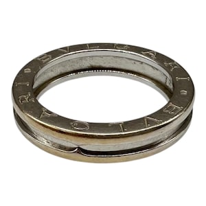 May include: A silver-toned metal ring featuring a smooth, rounded band with a central horizontal groove. The word BVLGARI is engraved in capital letters along the top surface of the band, encircling the entire circumference of the ring.