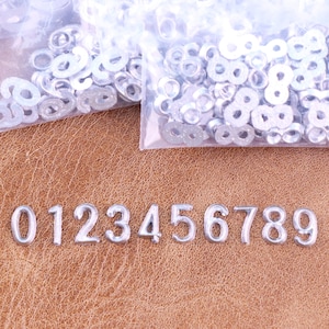 May include: Silver metal numbers 0 through 9 are displayed on a brown leather surface. Small silver metal eyelets are in plastic bags in the background. The numbers are in a serif font.