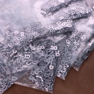 Może przedstawiać: Kilka przezroczystych plastikowych toreb wypełnionych małymi, srebrnymi metalowymi zawieszkami z literami i cyframi. Zawieszki są w różnych czcionkach i rozmiarach, idealne do tworzenia biżuterii lub projektów rzemieślniczych.