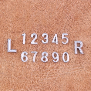 May include: Silver metal letters and numbers, including the letters "L" and "R", and the numbers 1 through 0, are arranged on a textured, brown leather surface. The numbers and letters are small and metallic.