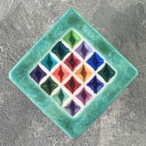 May include: A square ceramic tile with a diamond-shaped orientation, featuring a wide, mottled teal border. The center contains a grid of sixteen smaller, recessed diamond-shaped indentations, each glazed in a different vibrant color.