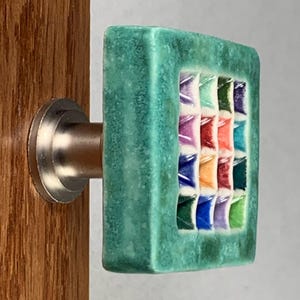 Pu&ograve; includere: Un pomello quadrato per mobile con una cornice testurizzata verde acqua e una griglia centrale di sedici piccoli quadrati incassati multicolori nelle tonalit&agrave; viola, blu, arancione, rosso e verde, fissato su una base in metallo argentato.