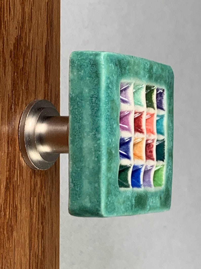 Pu&ograve; includere: Un pomello quadrato per mobile con una cornice testurizzata verde acqua e una griglia centrale di sedici piccoli quadrati incassati multicolori nelle tonalit&agrave; viola, blu, arancione, rosso e verde, fissato su una base in metallo argentato.