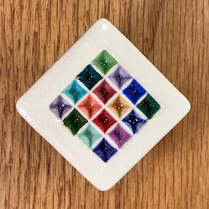 May include: A white, diamond-shaped ceramic tile with a colorful mosaic pattern. The mosaic features small, square tiles in shades of green, blue, purple, red, and yellow. The tile is set against a wooden background.