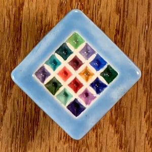 May include: A square, light blue ceramic tile with a diamond shape. The center features a grid of colorful squares in shades of green, red, purple, and yellow. The tile is set against a wooden background.