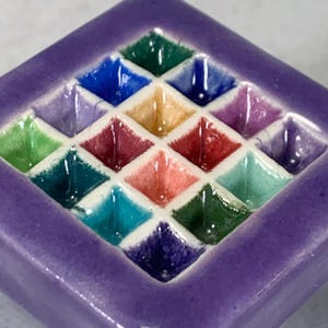 May include: A square ceramic tile featuring a grid of sixteen recessed, diamond-shaped compartments. Each compartment is glazed in a different vibrant color, including shades of blue, green, red, yellow, and purple, separated by white grout lines.