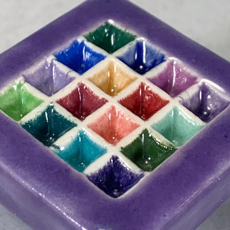 May include: A square ceramic tile featuring a grid of sixteen recessed, diamond-shaped compartments. Each compartment is glazed in a different vibrant color, including shades of blue, green, red, yellow, and purple, separated by white grout lines.