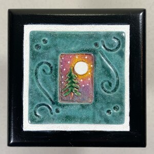 May include: A small wooden box with a teal blue ceramic tile inset. The tile features a whimsical design of a green tree with a white moon and pink and purple sky.