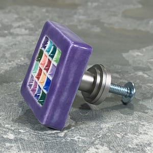 May include: A square purple ceramic cabinet knob featuring a central grid pattern of small, multi-colored squares in shades of blue, green, red, and orange. The knob includes a silver-toned metal base and a threaded screw for installation.