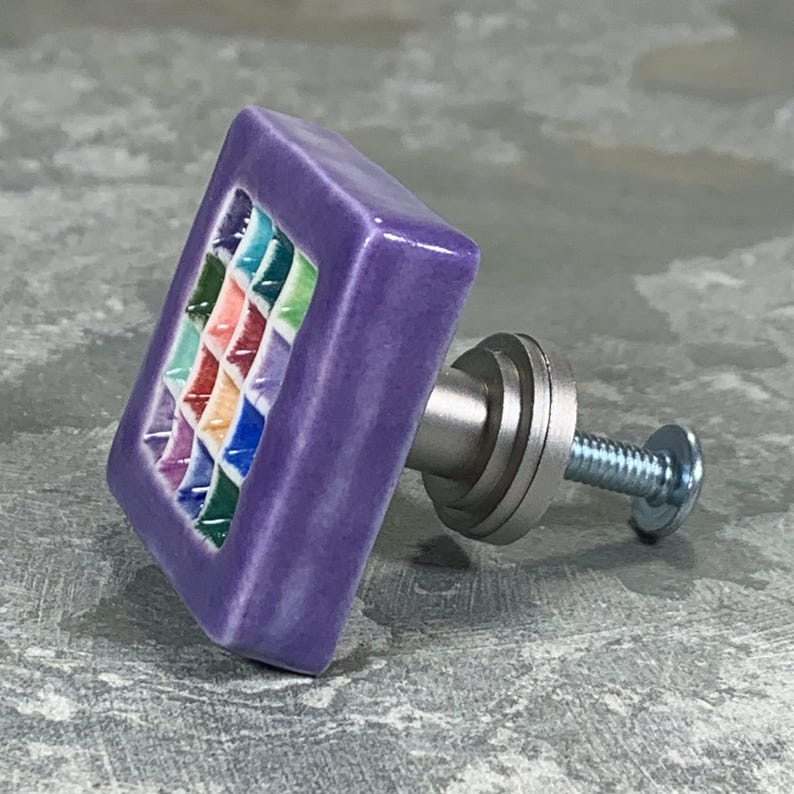 May include: A square purple ceramic cabinet knob featuring a central grid pattern of small, multi-colored squares in shades of blue, green, red, and orange. The knob includes a silver-toned metal base and a threaded screw for installation.