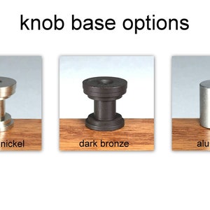 Puede incluir: Tres bases de pomo diferentes en n&iacute;quel cepillado, bronce oscuro y aluminio. Las bases se muestran sobre una superficie de madera.