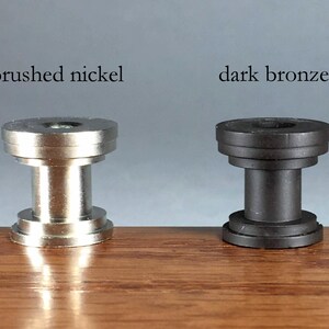 Pu&ograve; includere: Due componenti hardware cilindrici in metallo, uno con finitura in nichel spazzolato e uno in bronzo scuro. Ogni pezzo presenta una forma a bobina a gradini con parte superiore e inferiore piatte, etichettati come brushed nickel e dark bronze.